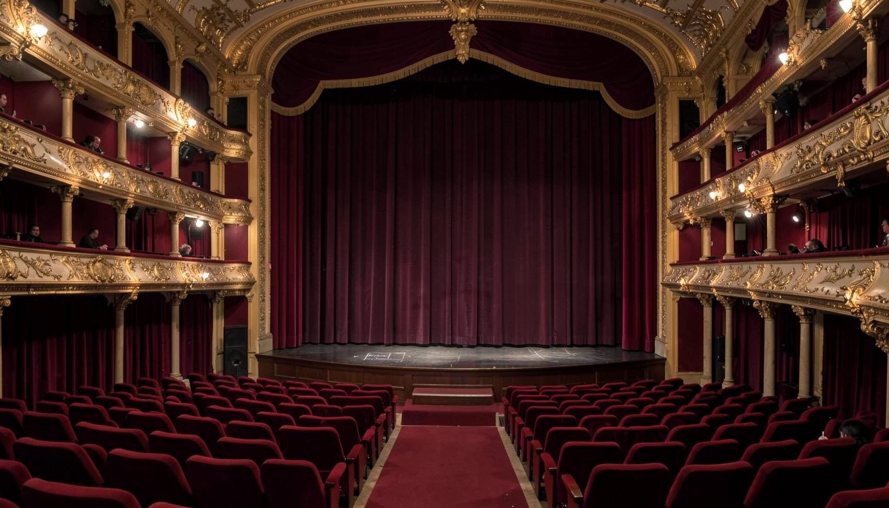 A grand Italian theater interior with rows of red velvet seats, ornate gilded balconies, and a softly lit stage framed by deep burgundy curtains