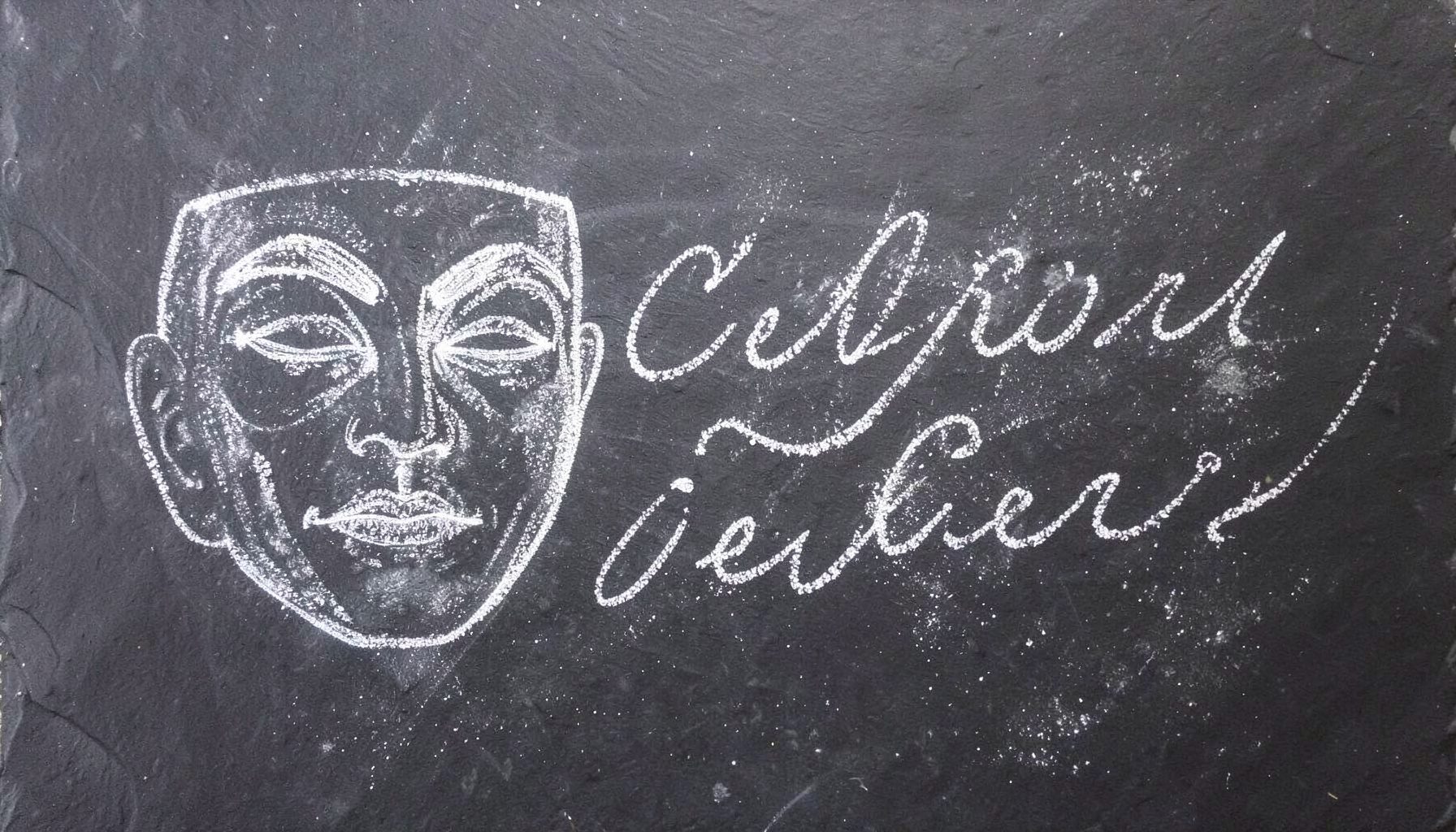 A chalk drawing on a dark slate board showing a theatrical mask alongside flowing script lettering, with soft smudges and chalk dust visible across the surface